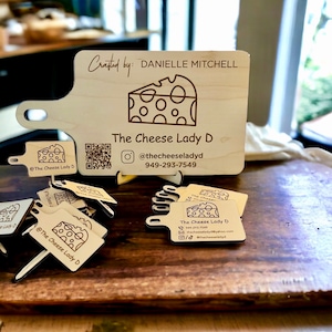May include: A wooden cheese board with a handle, featuring a laser-engraved design of a wedge of cheese. The board has the text "Crafted by: DANIELLE MITCHELL" and "The Cheese Lady D" with a QR code and social media information. There are also several smaller wooden cheese markers with the same design.