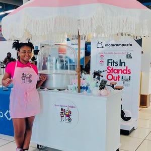 May include: A cotton candy cart with a pink and white striped umbrella and a white cart. The cart has the text "Street Carolina Delights" and a logo. A person is standing in front of the cart.