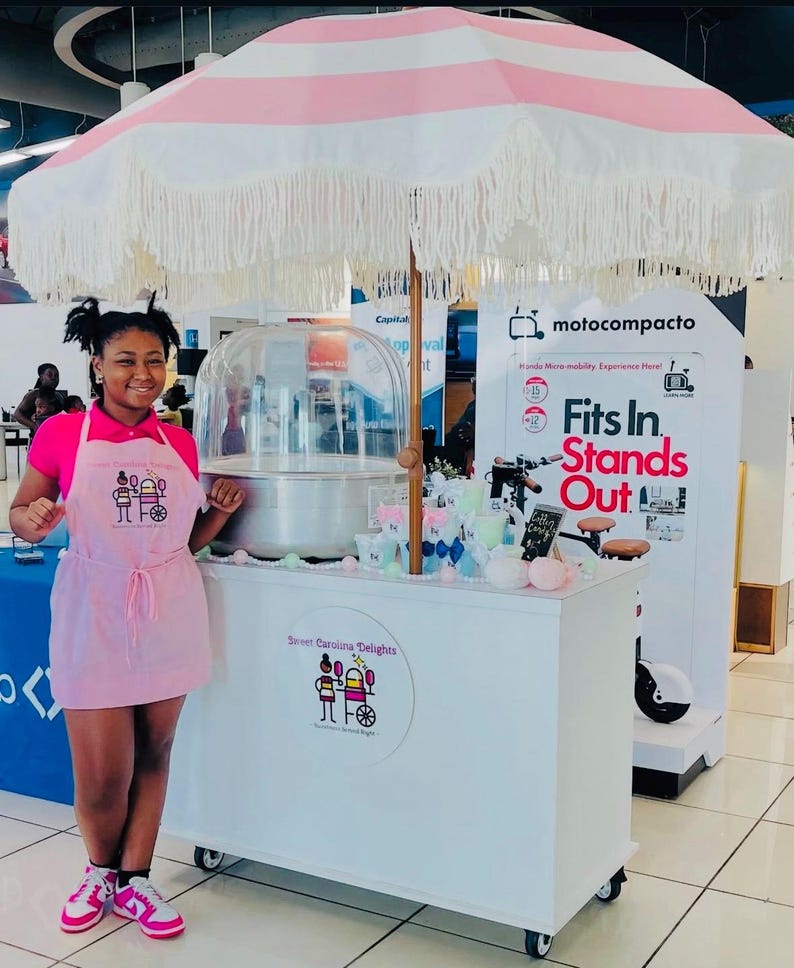 May include: A cotton candy cart with a pink and white striped umbrella and a white cart. The cart has the text "Street Carolina Delights" and a logo. A person is standing in front of the cart.