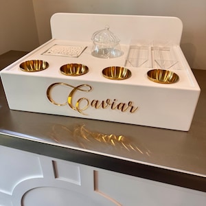 May include: A white caviar serving station with gold accents. It features four gold-colored serving dishes, a glass serving bowl, and clear acrylic containers. The word "Caviar" is displayed in gold script on the front.