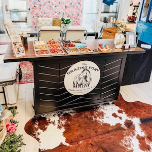 May include: A charcuterie bar with various food items displayed in metal trays. The bar features a black and brown design with the logo "The Grazing Pony." A cowhide rug is on the floor. The background includes a floral wall and a table with chairs.