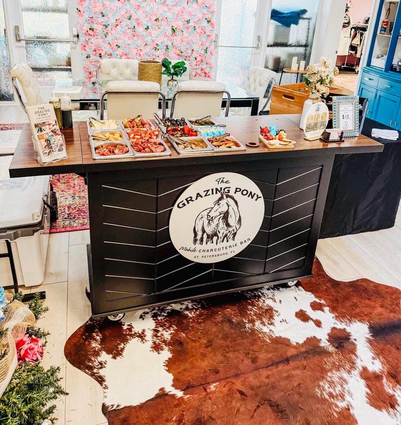 May include: A charcuterie bar with various food items displayed in metal trays. The bar features a black and brown design with the logo "The Grazing Pony." A cowhide rug is on the floor. The background includes a floral wall and a table with chairs.