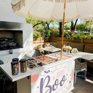 May include: A white outdoor cart with a Halloween theme, featuring the text "a little Boo is almost due." The cart has a white countertop with jars of candy and trays of treats. A white fringed umbrella provides shade.