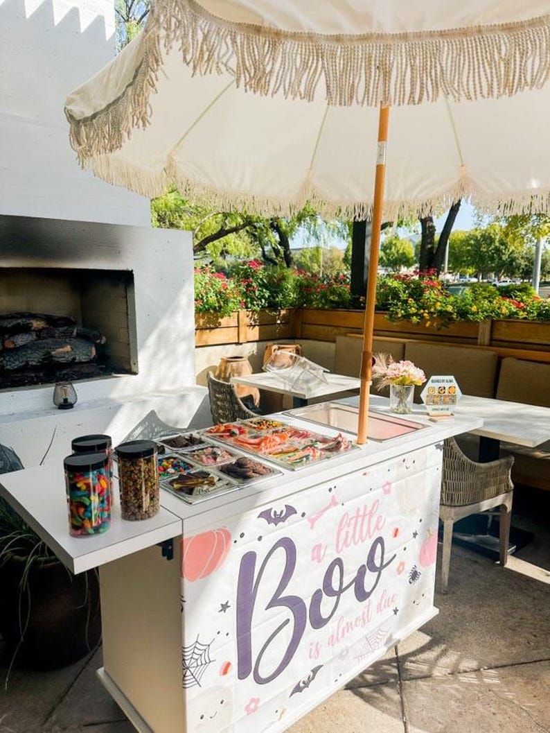 May include: A white outdoor cart with a Halloween theme, featuring the text "a little Boo is almost due." The cart has a white countertop with jars of candy and trays of treats. A white fringed umbrella provides shade.