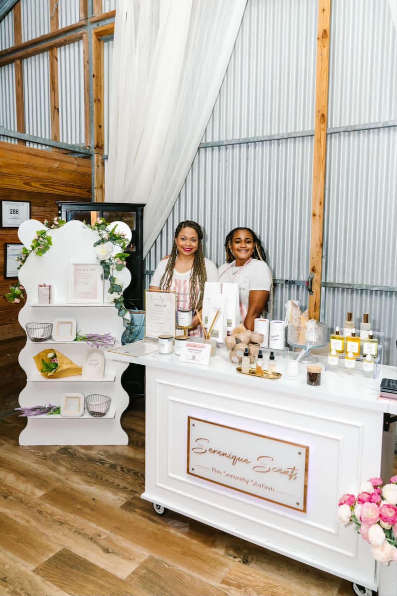 May include: Two women stand behind a white display table with products. The table has a sign that reads "Serenique Scents." A white shelving unit displays products and floral arrangements. The products include candles, oils, and other beauty items.