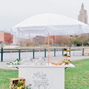 May include: A white charcuterie cart with a fringed umbrella, set up outdoors. The cart features a logo, clear domes, and a tiered serving tray. A wicker basket filled with lemons and greenery sits nearby.