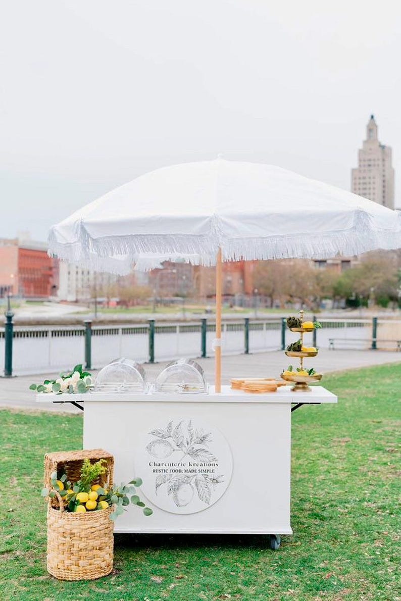 May include: A white charcuterie cart with a fringed umbrella, set up outdoors. The cart features a logo, clear domes, and a tiered serving tray. A wicker basket filled with lemons and greenery sits nearby.