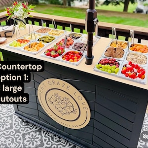 May include: A charcuterie bar with a black base and a light wood countertop. The countertop holds various food items in stainless steel trays. The bar features the text "Eat Graze Love Charcuterie Bar" on a wooden sign. The text "Countertop option 1: 3 large cutouts" is also visible.