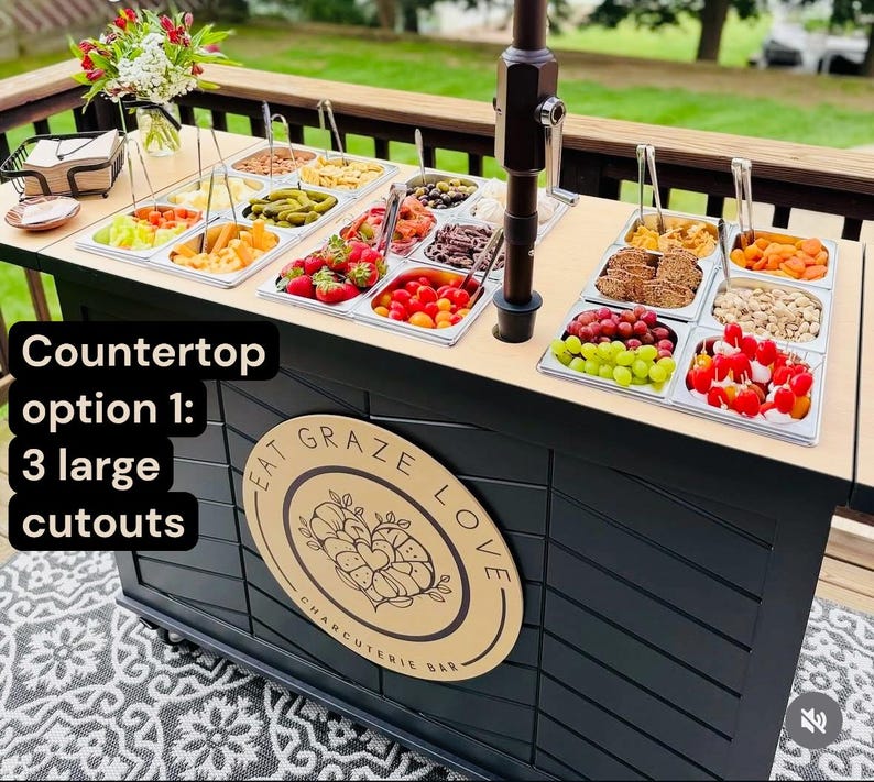 May include: A charcuterie bar with a black base and a light wood countertop. The countertop holds various food items in stainless steel trays. The bar features the text "Eat Graze Love Charcuterie Bar" on a wooden sign. The text "Countertop option 1: 3 large cutouts" is also visible.