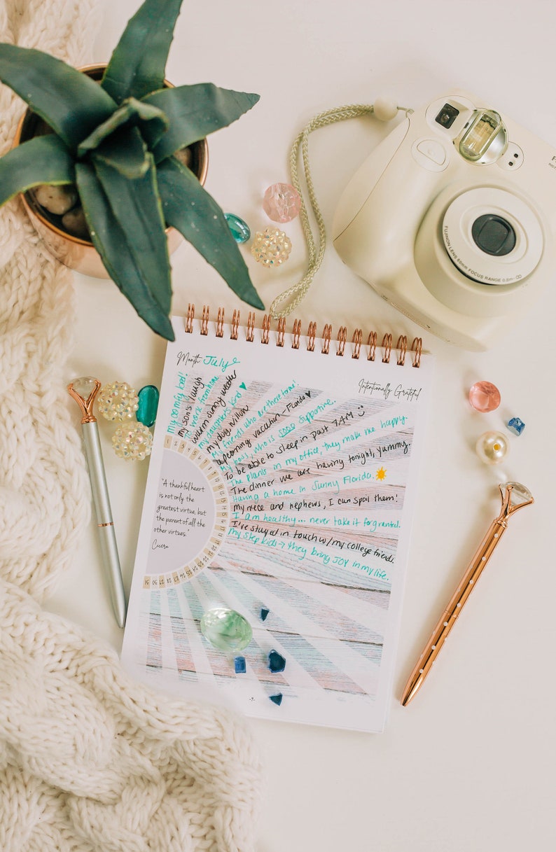 Puede incluir: Un cuaderno blanco de espiral con un fondo colorido y texto escrito a mano. El texto incluye una lista de cosas por las que el escritor est&aacute; agradecido, como "&iexcl;mi cama c&oacute;moda!" y "la risa de mi hijo". El cuaderno est&aacute; rodeado de una manta de punto blanco, una planta suculenta en maceta, una c&aacute;mara instant&aacute;nea rosa y blanca y gemas de vidrio de colores.
