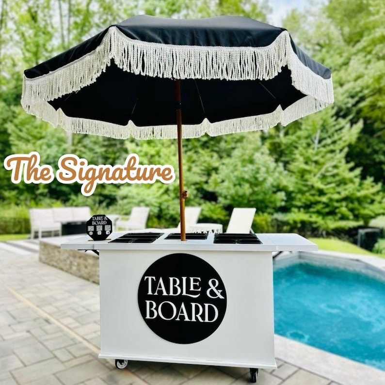 May include: A white mobile bar with a black umbrella featuring white fringe. The bar has a black and white logo that says "TABLE & BOARD". The text "The Signature" is also visible.