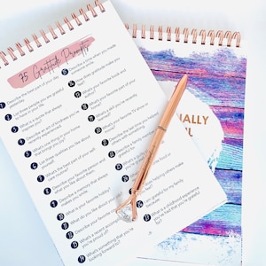 Puede incluir: Un cuaderno espiralado con una portada rosa y blanca que dice "75 Gratitude Prompts". El cuaderno est&aacute; abierto en una p&aacute;gina con una lista de indicaciones para practicar la gratitud. Un bol&iacute;grafo de color rosa dorado est&aacute; sobre el cuaderno.