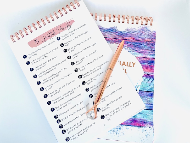 Puede incluir: Un cuaderno espiralado con una portada rosa y blanca que dice "75 Gratitude Prompts". El cuaderno est&aacute; abierto en una p&aacute;gina con una lista de indicaciones para practicar la gratitud. Un bol&iacute;grafo de color rosa dorado est&aacute; sobre el cuaderno.