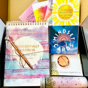 Puede incluir: Una caja negra con una variedad de art&iacute;culos de cuidado personal, que incluyen una libreta rosa y azul con las palabras "Intentionally Grateful" en la portada, un bol&iacute;grafo dorado, un juego de cartas azul y blanco con las palabras "Mindset Cards", una peque&ntilde;a bomba de ba&ntilde;o rosa y naranja, y una peque&ntilde;a tarjeta con las palabras "Hand up, gorgeous. Straighten that crown."