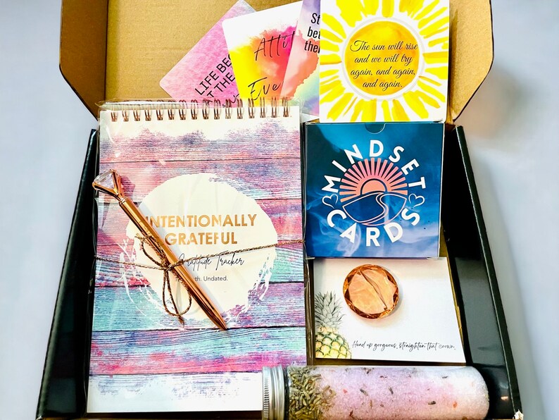 Puede incluir: Una caja negra con una variedad de art&iacute;culos de cuidado personal, que incluyen una libreta rosa y azul con las palabras "Intentionally Grateful" en la portada, un bol&iacute;grafo dorado, un juego de cartas azul y blanco con las palabras "Mindset Cards", una peque&ntilde;a bomba de ba&ntilde;o rosa y naranja, y una peque&ntilde;a tarjeta con las palabras "Hand up, gorgeous. Straighten that crown."
