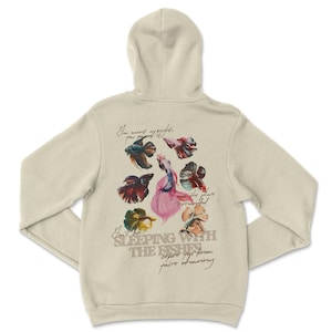 May include: Cream-coloured hoodie with a hood. The back features a colourful illustration of several betta fish and handwritten text that reads "You want of nights, you pound, the place surrounded, You'll be sleeping with the fishes before you know you're drowning."