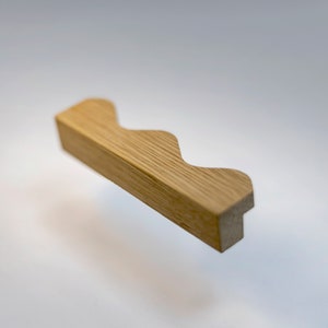May include: A wooden drawer pull with a wavy, undulating design. The pull is light brown in colour and has a smooth, polished finish.