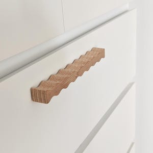 May include: A light brown wooden drawer pull with a distinct wavy, undulating shape and a visible natural wood grain texture. The handle is mounted horizontally onto a smooth, matte white cabinet drawer, creating a contrast in color and material.