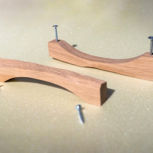 Könnte beinhalten: Zwei Holzknaufgriffe für Schranktüren mit gebogenem Design. Die Griffe sind aus hellem braunem Holz gefertigt und haben eine glatte Oberfläche. Jeder Griff hat eine silberne Schraube in der Mitte.