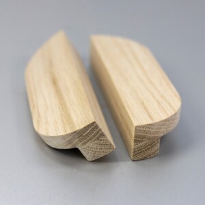 Puede incluir: Dos tiradores de armario de madera de color claro con un patrón de veta natural. Cada tirador presenta una superficie superior lisa y curva y una base rectangular, mostrando la textura distintiva y el color tostado claro de la madera.