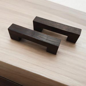 Peut inclure: Deux poignées de tiroir en bois brun foncé de forme rectangulaire avec une poignée découpée.