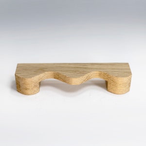 May include: A wooden shelf with a wavy edge and two legs. The shelf is made of light brown wood and has a natural finish.