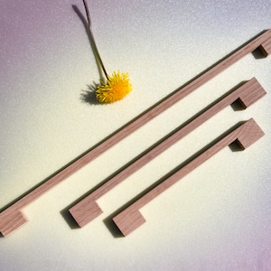 May include: Three wooden cabinet pulls in various sizes, arranged diagonally on a light grey background. The pulls are made of natural wood and have a simple, rectangular design.