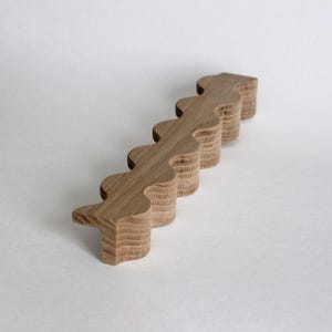 May include: A light brown wooden handle with a wavy, scalloped edge design. The object features a layered, stacked construction showing the natural wood grain texture along its sides, with a repeating curved pattern along the front edge.