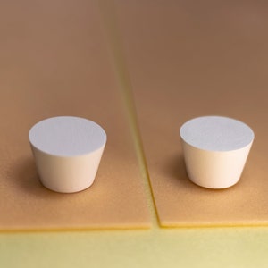 May include: Two white, truncated cone-shaped objects with flat circular tops and smooth, matte surfaces. These small, solid items feature a tapered design, narrowing from the top toward the base, and are presented in a side-by-side arrangement.