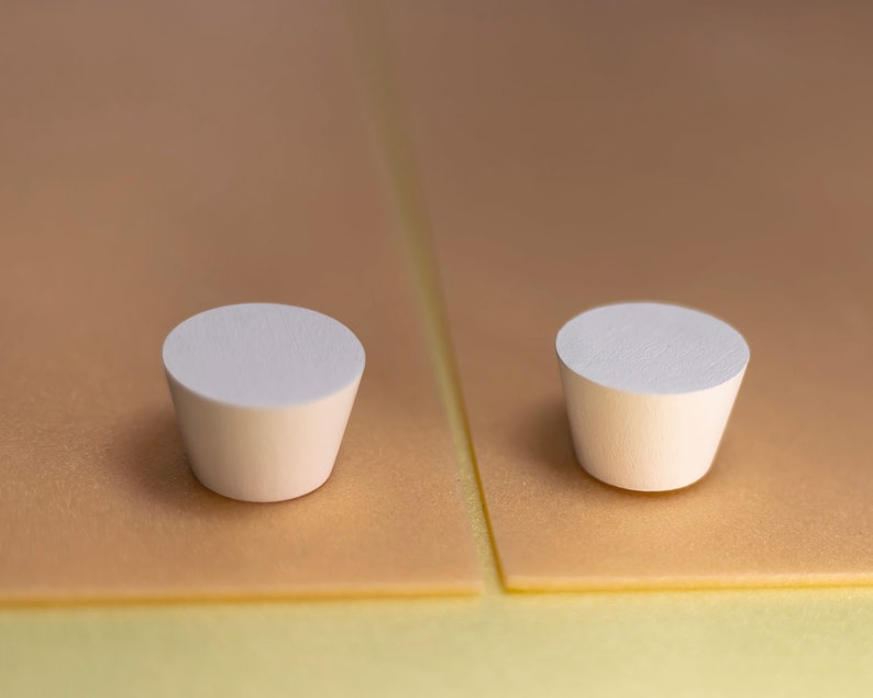 May include: Two white, truncated cone-shaped objects with flat circular tops and smooth, matte surfaces. These small, solid items feature a tapered design, narrowing from the top toward the base, and are presented in a side-by-side arrangement.