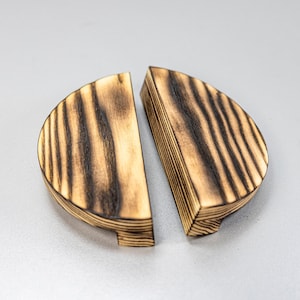May include: Two semi-circular wooden cabinet pulls featuring a light tan natural wood grain with dark, charred vertical stripes. Each piece has a flat straight edge and a curved outer edge, showcasing a distinct scorched wood finish throughout.