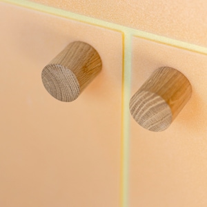 May include: Two wooden knobs with a light brown finish are mounted on a peach-colored surface. The knobs are cylindrical in shape and have a slightly textured surface.