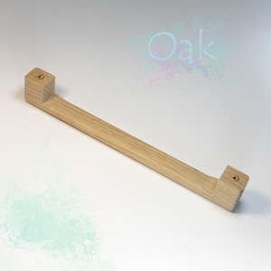 Puede incluir: Un tirador de cajón de madera largo, de color marrón claro, con forma cuadrada en cada extremo. El tirador tiene un diseño sencillo y está hecho de madera natural.