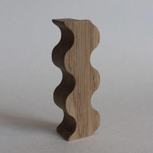 May include: A vertical wooden object with a wavy, undulating silhouette on one side and a flat, straight edge on the other. The piece features a natural light brown wood grain pattern with visible vertical lines running throughout the entire structure.