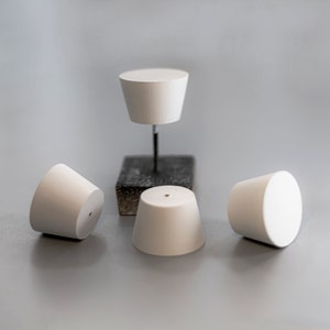 May include: Four white, truncated cone-shaped stoppers with smooth surfaces. Three stoppers rest on the surface, while one is elevated on a thin metal rod attached to a small, dark, square-shaped base. Each stopper features a small, centered hole.