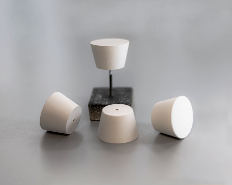 May include: Four white, truncated cone-shaped stoppers with smooth surfaces. Three stoppers rest on the surface, while one is elevated on a thin metal rod attached to a small, dark, square-shaped base. Each stopper features a small, centered hole.