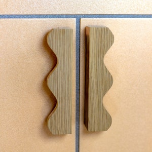 May include: Two wooden cabinet door pulls with a wavy edge design. The pulls are a light brown colour and are mounted on a light brown tiled surface.