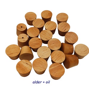 May include: A collection of twenty-one light brown, tapered wooden bottle stoppers with a smooth, oiled finish. Each stopper has a circular top and a conical body. Two of the stoppers feature a small, centered hole. The text "alder + oil" is visible.