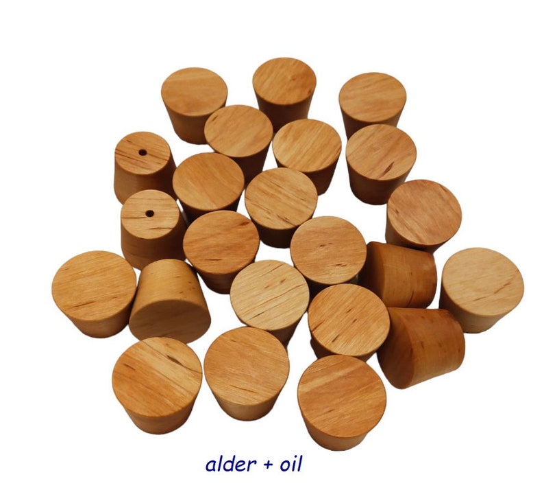 May include: A collection of twenty-one light brown, tapered wooden bottle stoppers with a smooth, oiled finish. Each stopper has a circular top and a conical body. Two of the stoppers feature a small, centered hole. The text "alder + oil" is visible.