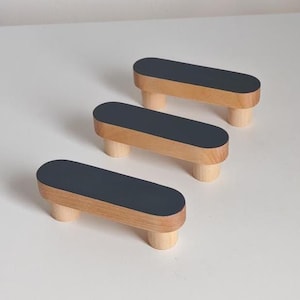 Puede incluir: Tres tiradores de armario de madera con una parte superior gris oscuro y una base de madera natural. Los tiradores tienen forma rectangular redondeada y están elevados por tres patas cilíndricas. Los tiradores están dispuestos en diagonal.