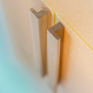 May include: Two wooden cabinet door pulls with a simple, rectangular design. The pulls are mounted on a light orange surface.