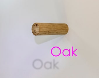 Może przedstawiać: Cylindryczny haczyk ścienny z jasnego drewna dębowego z widocznym wzorem naturalnych słojów na okrągłym końcu. Słowo Oak jest wydrukowane jaskraworóżowym tekstem pod haczykiem, z bladym szarym cieniem słowa Oak poniżej.