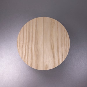 Puede incluir: Un disco redondo de madera con un acabado de grano de madera natural. El disco está hecho de dos piezas de madera que se unen.