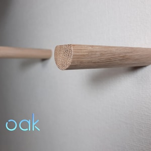 Può includere: Due ganci da parete in legno di quercia. I ganci sono arrotondati alle estremità e sono montati su una parete bianca. La parola "quercia" è scritta in blu sotto i ganci.