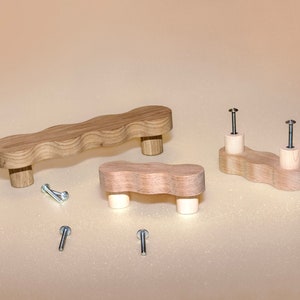 May include: Three wooden cabinet knobs with a wavy design. The knobs are made of light wood and have a natural finish. The knobs are shown with screws.