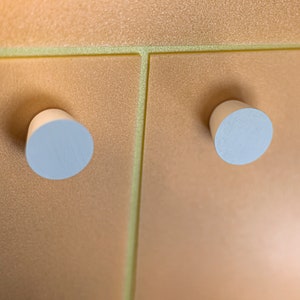 May include: Two cylindrical, light-colored wooden knobs are attached to a metallic, shimmering gold-toned surface. The surface features a distinct vertical seam and a horizontal seam, creating a grid-like pattern across the textured, reflective panel.