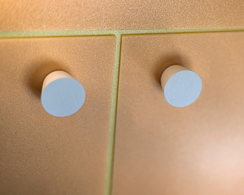 May include: Two cylindrical, light-colored wooden knobs are attached to a metallic, shimmering gold-toned surface. The surface features a distinct vertical seam and a horizontal seam, creating a grid-like pattern across the textured, reflective panel.