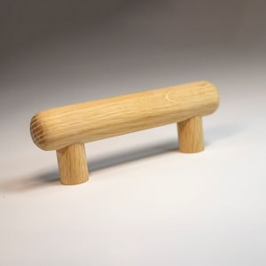 Puede incluir: Un tirador de cajón de madera con forma cilíndrica y bordes redondeados. El tirador tiene dos soportes cilíndricos cortos en cada extremo.