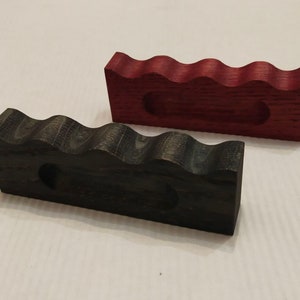 May include: Two wooden pipe stands. One is stained a dark ebony color, and the other is a deep red. Both have a wavy top with four curved grooves to hold pipes. Each stand has a circular hole.