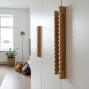 May include: Two vertical light brown wooden cabinet handles with a distinct wavy, scalloped edge design mounted on a smooth white door surface. The handles feature a long, narrow recessed grip area along the side for functional opening.